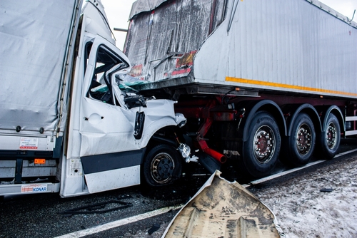 A significant collision involving two trucks is shown. The front of a white delivery truck is severely damaged after crashing into the back of a larger trailer truck. The road appears wet, suggesting slippery conditions, with visible debris scattered from the crash.